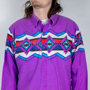 May include: A purple long-sleeved button-down shirt with a geometric pattern in shades of blue, red, and white. The shirt has a pointed collar and a single chest pocket.
