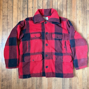 May include: A red and black buffalo plaid jacket featuring a wide collar, long sleeves, and a front button closure. The garment includes two chest pockets with flaps and two lower pockets with flaps, all secured with dark buttons.