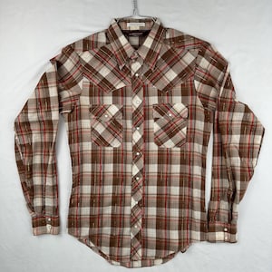 May include: A long-sleeved shirt in a brown, red, and cream plaid pattern, featuring two chest pockets and pearl snap closures. The shirt is suspended from a metal hanger. It has a classic western design.