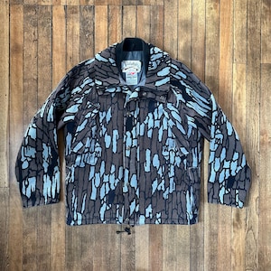 May include: A brown and blue camouflage pattern jacket with a black collar and a drawstring at the waist. The jacket has a button-up front and multiple pockets. The brand name "Cabela's" is visible on a tag near the collar.