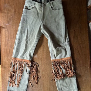 May include: Green suede pants with brown fringe trim at the bottom. The pants have suspenders attached.