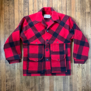 May include: A long-sleeve button-up jacket featuring a bold red and black buffalo plaid pattern. The garment includes a pointed collar, two large front patch pockets with flaps, and a central button closure. The fabric has a textured wool-like finish.