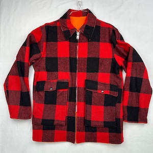 May include: A red and black checked jacket with a full zip and two front pockets. The jacket has a black collar and an orange lining. The jacket is suspended from a metal hanger.
