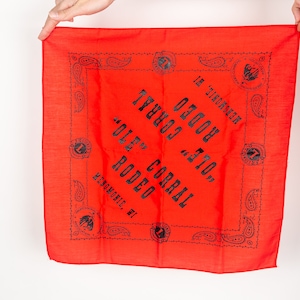 Op de afbeelding: Rode bandana met een zwarte paisley-rand en de tekst "OLE' CORRAL RODEO MENOMONIE, WI" in het zwart gedrukt.