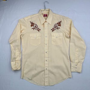 May include: A long-sleeved, light yellow button-down shirt with two chest pockets and pearl snap closures. The shirt features brown embroidered designs on the chest. The shirt is hanging on a metal hanger.