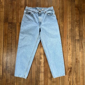 Könnte beinhalten: Hellblaue Jeans mit klassischem Fünf-Taschen-Design. Die Jeans haben einen geraden Beinverlauf und einen Knopf- und Reißverschluss. Der Denim scheint ein mittleres Gewicht zu haben. Die Jeans liegen flach auf einer Holzoberfläche.