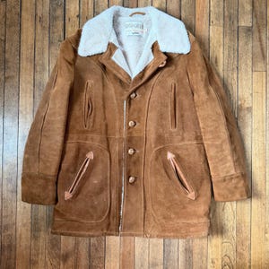 Könnte beinhalten: Ein brauner Wildledermantel mit cremefarbenem Shearling-Kragen und -Futter. Der Mantel hat vier Knöpfe, zwei Vordertaschen und zwei Brusttaschen. Der Mantel ist im Vintage-Stil.