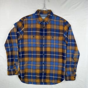 Puede incluir: Una camisa de cuadros de manga larga en tonos azules, dorados y marrones, con dos bolsillos en el pecho y cierres de botones. La camisa tiene el texto "1969" en el cuello.