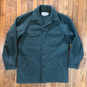 May include: A dark forest green wool-blend jacket featuring a collared neckline, a button-down front, and long sleeves with buttoned cuffs. The garment includes two chest pockets with buttoned flaps and two lower slit pockets, with a white label inside.