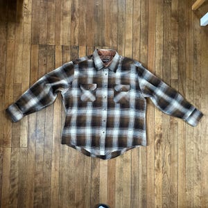 Può includere: Una camicia di flanella a quadri marrone, bianca e nera con abbottonatura, due tasche sul petto e colletto marrone. La camicia a maniche lunghe è stesa su una superficie di legno.