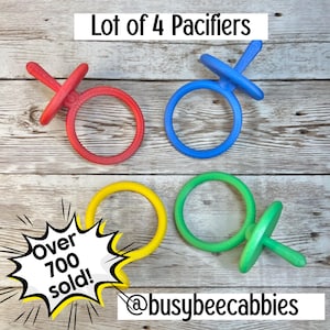 Op de afbeelding: Een set van 4 fopspenen in rood, blauw, geel en groen. De fopspenen hebben een ronde ring en een centrale steel. De tekst op de afbeelding luidt: Lot of 4 Pacifiers, Over 700 sold! en @busybeecabbies, geplaatst op een houten ondergrond.