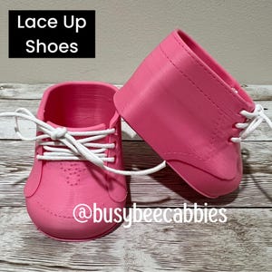 May include: A pair of bright pink 3D printed shoes featuring white laces and a lace-up design. The text "Lace Up Shoes" appears in a black box in the upper left corner, and the text "@busybeecabbies" is printed in white at the bottom centre.
