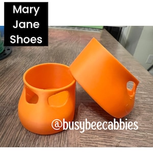 May include: Two bright orange, cup-shaped silicone protective boots with a cutout design. The text "Mary Jane Shoes" appears in a black box at the top, and "@busybeecabbies" is printed in white at the bottom of the image.