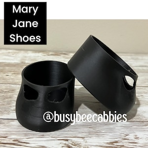May include: Two black, cylindrical, 3D-printed shoe covers featuring a cutout design on the sides. The image includes the text "Mary Jane Shoes" in a black box and the social media handle "@busybeecabbies" printed in white text.