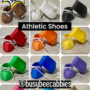 Könnte beinhalten: Eine Sammlung von Miniatur-Sportschuhen für Cabbage Patch Kids Puppen in verschiedenen Farben wie Braun, Weiß, Schwarz, Rot, Orange, Gelb, Grün, Blau, Lila, Pink, Türkis und Beige. Jeder Schuh hat weiße Schnürsenkel und Textüberlagerungen.
