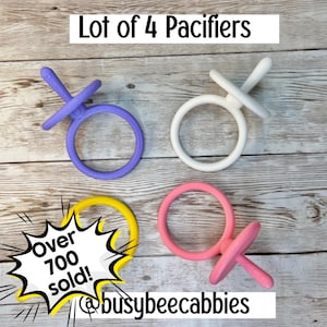 May include: A lot of 4 pacifiers in purple, white, yellow, and pink. The text Lot of 4 Pacifiers is at the top. A graphic burst with the text Over 700 sold! is on the left. The text busybeecabbies is at the bottom of this set of four pacifiers.