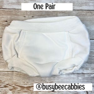 Può includere: Un paio di copripannolini bianco tinta unita in tessuto testurizzato, con un'ampia fascia elastica in vita e aperture elasticizzate per le gambe. L'immagine include il testo One Pair e l'handle @busybeecabbies stampati sul display.