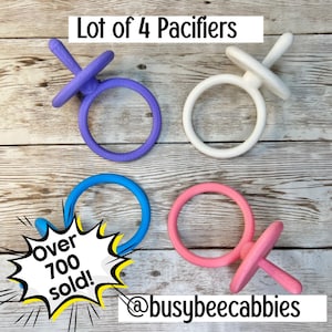 Op de afbeelding: Een set van vier fopspenen in paars, wit, blauw en roze. Elke fopspeen heeft een ronde ringgreep en een centrale speen. De afbeelding bevat de tekst Lot of 4 Pacifiers, Over 700 sold!, en @busybeecabbies.