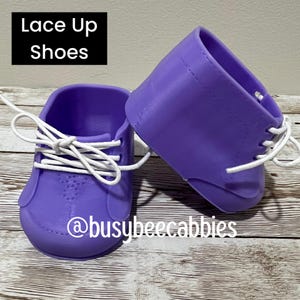 May include: A pair of purple doll shoes with white laces, featuring the text Lace Up Shoes and @busybeecabbies. The shoes have a smooth, moulded finish with visible stitching details around the ankle collar and eyelets for the white laces.
