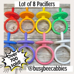 May include: A lot of 8 pacifiers in red, orange, yellow, green, blue, purple, pink, and white. Each pacifier features a round shield with a handle ring. The text Lot of 8 Pacifiers, Over 700 sold!, and @busybeecabbies is visible on the image.
