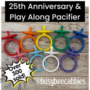 Può includere: Una collezione di nove giocattoli in silicone a forma di ciuccio in rosso, arancione, giallo, verde, blu, rosa, azzurro, bianco e viola. Il testo recita 25th Anniversary & Play Along Pacifier e Over 300 sold! con @busybeecabbies.