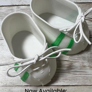 May include: A pair of white shoes with bright green accents on the sides and white laces tied in bows. The shoes feature a smooth, molded construction with a rounded toe design. The text "Now Available: Two-Color Shoes" is printed at the bottom.