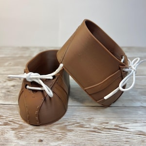 May include: A pair of brown shoes with a smooth, matte finish and white laces tied in bows. The shoes feature a rounded toe design with visible stitched detailing along the front and sides, creating a structured look for the footwear.