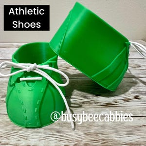 Puede incluir: Dos recipientes de color verde brillante impresos en 3D con forma de calzado deportivo, con cordones blancos pasados por ojales. El texto "Athletic Shoes" aparece en un recuadro negro y "@busybeecabbies" está impreso en blanco abajo a la derecha.
