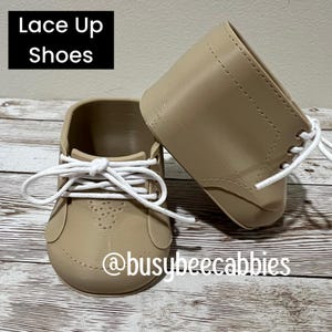 May include: A pair of beige shoes with white laces and a lace-up design. The text "Lace Up Shoes" appears in a black box in the upper left corner, and the text "@busybeecabbies" is printed in white across the lower portion of the shoes.