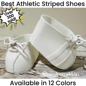 May include: Two white shoe-shaped containers with white laces, featuring the text Best Athletic Striped Shoes, Over 300 sold!, and Available in 12 Colours. The objects have a smooth finish and are positioned to show the lacing and side details.