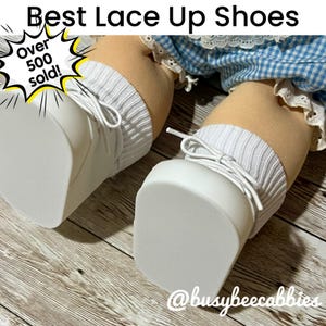 May include: A pair of white lace-up shoes worn by a doll with tan skin, featuring white ribbed socks and blue and white checkered fabric trim. The image includes the text Best Lace Up Shoes, Over 500 sold!, and @busybeecabbies.