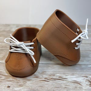 May include: A pair of brown doll shoes featuring a smooth finish, rounded toes, and white laces threaded through three pairs of eyelets on each side. The shoes have a structured design with visible stitching details along the sides and toe cap.