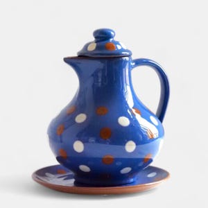 Könnte beinhalten: Eine blaue Keramikkanne mit weißen und braunen Punkten und einer passenden Untertasse. Die Kanne hat einen Deckel und einen Henkel.