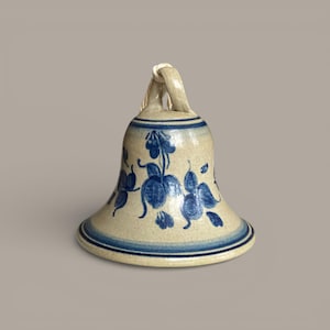 May include: A ceramic bell with a beige base and blue floral designs. The bell has a flared base with blue stripes and a loop at the top for hanging. The floral pattern includes leaves and flowers.