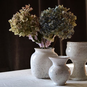 May include: Three speckled white ceramic vases of varying sizes, holding dried hydrangeas with green, blue, and purple hues. The vases are on a white surface against a dark brown backdrop.