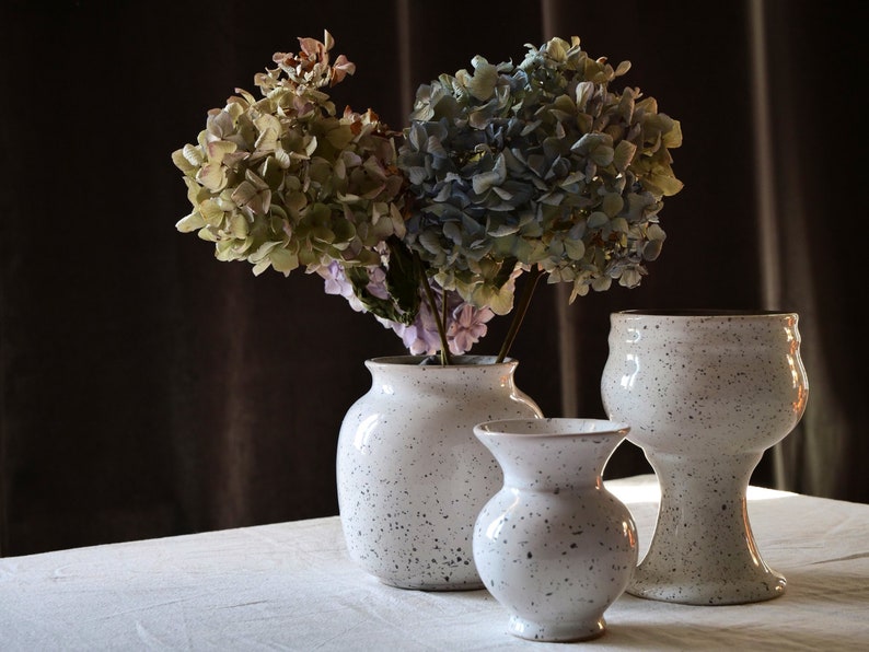 May include: Three speckled white ceramic vases of varying sizes, holding dried hydrangeas with green, blue, and purple hues. The vases are on a white surface against a dark brown backdrop.