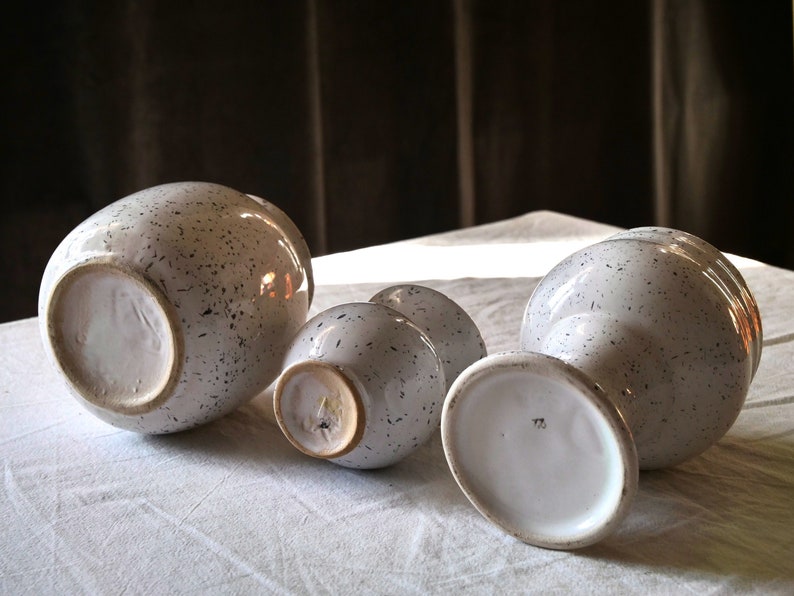 May include: A collection of white ceramic vases and bowls with a speckled black pattern. The pieces vary in size and shape, with some having a rounded form and others a more elongated design. The objects are arranged on a white surface.