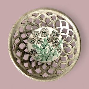 Pode incluir: Uma tigela decorativa vazada em tons de prata com um design floral central. A tigela apresenta um formato circular com um padrão de treliça ao redor da borda. O centro tem uma inserção de cerâmica branca com um motivo floral verde e marrom.
