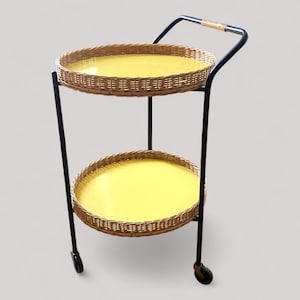 May include: A two-tiered serving cart with a black metal frame. The round trays have woven wicker rims and bright yellow centers. The handle is wrapped in woven material. The cart has small wheels.