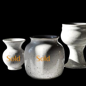 May include: Three speckled white ceramic vases of varying shapes. The vases have a matte finish and a dark rim. The word "Sold" is printed on two of the vases. The background is black.