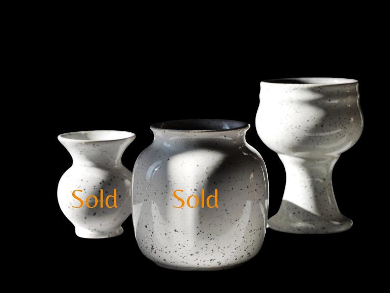 May include: Three speckled white ceramic vases of varying shapes. The vases have a matte finish and a dark rim. The word "Sold" is printed on two of the vases. The background is black.