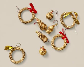 May include: A collection of gold-colored straw Christmas ornaments. The ornaments include wreaths with red and gold ribbons, teardrop shapes, and round ball shapes. The ornaments are arranged on a neutral background.