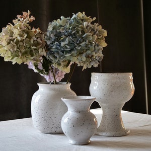 May include: Three white ceramic vases with a speckled design, holding dried hydrangeas in shades of green, blue, and purple. The vases vary in size and shape, creating a decorative display.