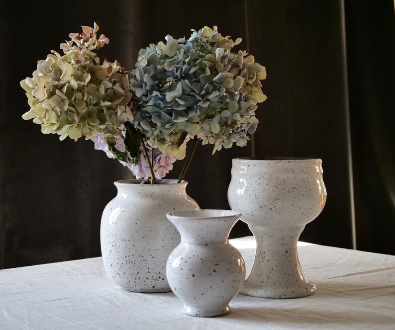 May include: Three white ceramic vases with a speckled design, holding dried hydrangeas in shades of green, blue, and purple. The vases vary in size and shape, creating a decorative display.