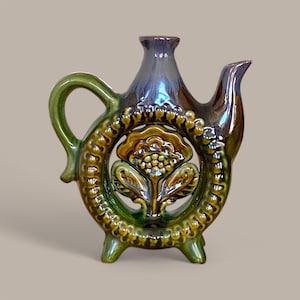May include: A green and brown ceramic teapot with a floral design and a cutout in the shape of a flower. The teapot has a handle and a spout and sits on three legs.