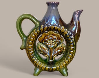 May include: A green and brown ceramic teapot with a floral design and a cutout in the shape of a flower. The teapot has a handle and a spout and sits on three legs.