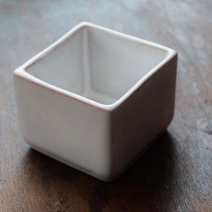 Peut inclure: Un petit récipient carré en céramique blanche avec une surface légèrement texturée. Le bord a un subtil accent marron. Le récipient est vide et repose sur une surface en bois sombre. Idéal pour les petites plantes ou comme pièce décorative.