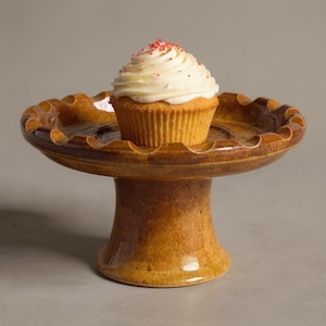 Könnte beinhalten: Ein brauner Keramik-Kuchenständer mit gewelltem Rand, auf dem ein Cupcake mit weißem Zuckerguss und roten Streuseln steht. Der Ständer hat einen Sockel. Der Ständer ist in Braun- und Beigetönen glasiert.