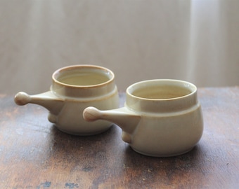 May include: Two beige ceramic soup bowls with handles. The bowls have a rounded shape with a small handle on one side and a slightly flared rim. The interior is a lighter shade of beige, and the rims have a brown accent.
