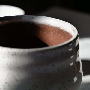 May include: Close-up of a speckled white ceramic mug with a textured exterior. The interior is a warm brown color. The rim has a white and dark speckled pattern. The mug is partially in shadow.